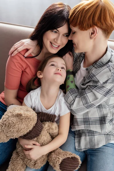 Sorridente madri abbracciando vicino figlia con orsacchiotto sul divano di casa — Foto stock