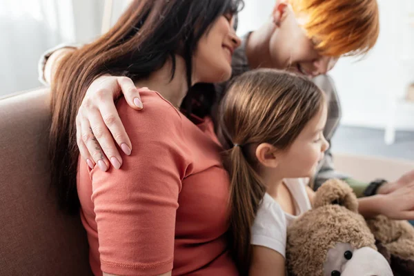 Seitenansicht von lächelnden gleichgeschlechtlichen Eltern, die mit Tochter sitzen, die Teddybär auf Couch hält — Stockfoto