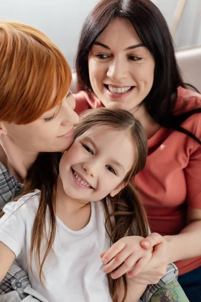 Cheerful kid smiling at camera near same sex parents on couch — Stock Photo