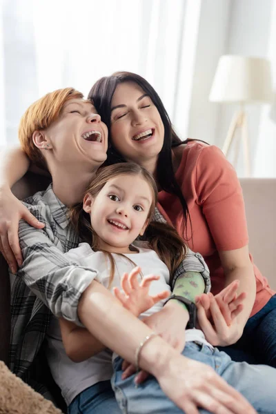 Foco seletivo dos pais do mesmo sexo rindo ao abraçar a filha no sofá na sala de estar — Fotografia de Stock