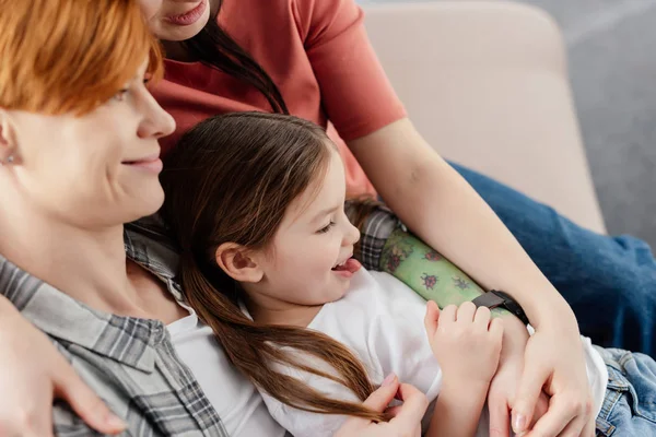 Seitenansicht eines lächelnden gleichgeschlechtlichen Paares, das seine Tochter auf der Couch umarmt — Stockfoto