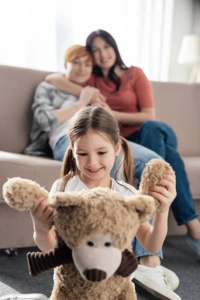 Selektiver Fokus eines lächelnden Kindes, das mit Teddybär in der Nähe von Müttern spielt, die sich auf der Couch umarmen — Stockfoto