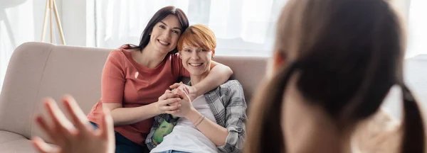 Selective focus of smiling mothers looking at daughter waving hand at home, panoramic shot — Stock Photo