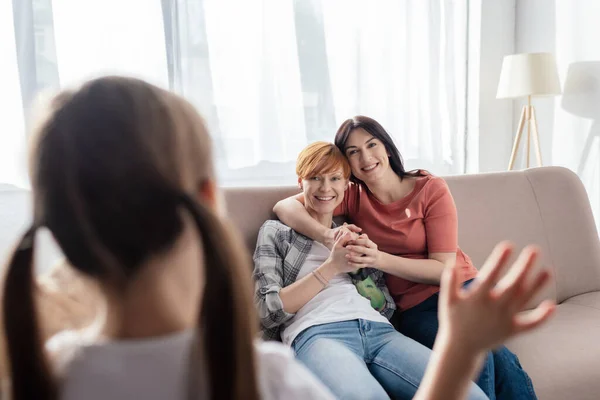 Selective focus of smiling same sex parents looking at daughter waving hand at home — Stock Photo