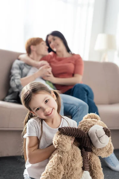 Selective focus of child with teddy bear smiling at camera near same sex parents embracing on couch at home — Stock Photo