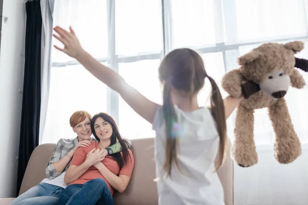 Selective focus of smiling mothers looking at daughter with teddy bear in living room — Stock Photo