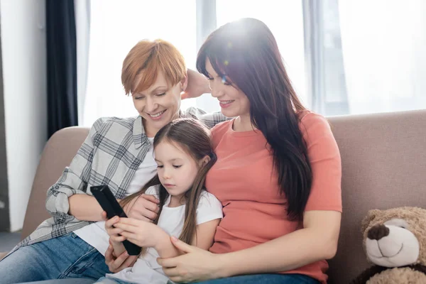 Mères souriantes assis près de la fille avec télécommande sur le canapé — Photo de stock