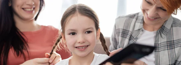 Colpo panoramico di madri sorridenti sedute vicino a bambino allegro con telecomando sul divano — Foto stock