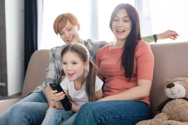 Fröhliches Kind mit Fernbedienung sitzt neben Müttern auf Couch im Wohnzimmer — Stockfoto