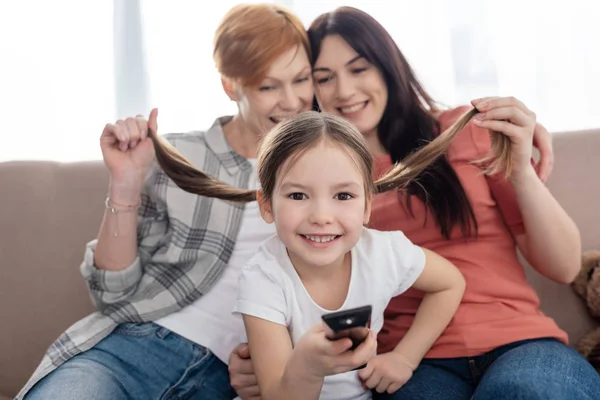 Selective focus of cheerful kid with remote controller smiling at camera near mothers holding hair on couch — Stock Photo