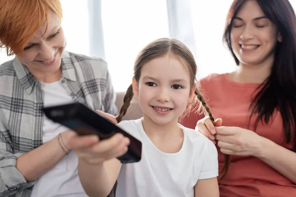 Selective focus of happy kid with remote controller near mothers plaiting pigtails on couch — Stock Photo