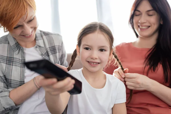 Focus selettivo del bambino felice con telecomando vicino alle madri che intrecciano trecce sul divano — Foto stock