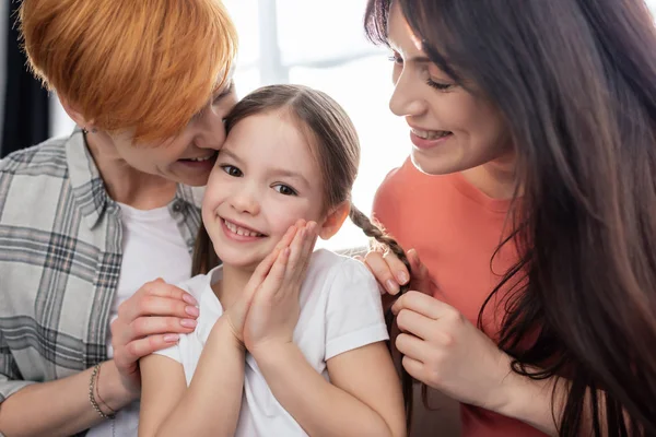 Lächelndes Kind, das in die Kamera schaut, in der Nähe glücklicher gleichgeschlechtlicher Eltern zu Hause — Stockfoto