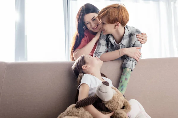 Smiling mothers embracing and looking at daughter with teddy bear on couch at home — Stock Photo