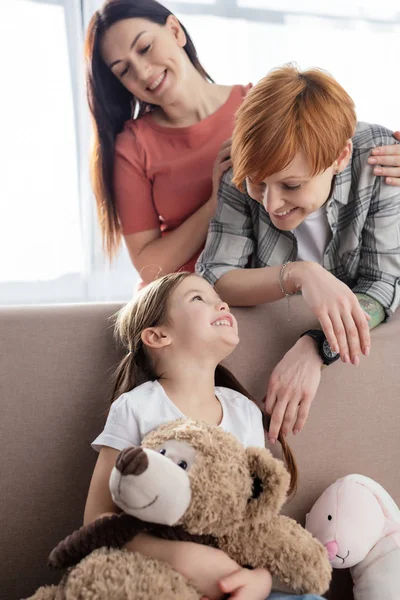 Selective focus of cheerful kid with soft toys on couch looking at same sex parents in living room — Stock Photo