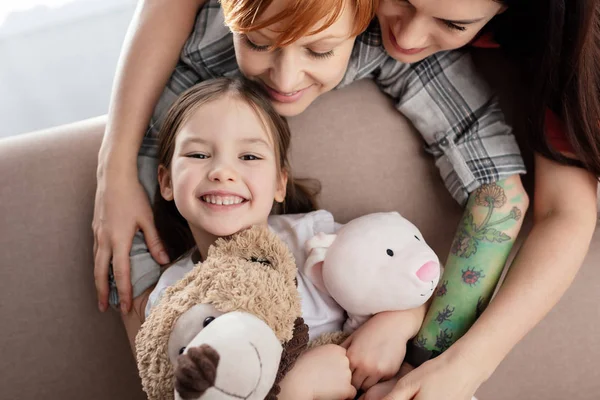 Smiling mothers hugging happy child with soft toys looking at camera on couch — Stock Photo