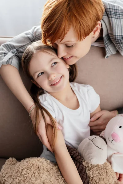 Sorrindo mãe abraçando filha com brinquedos macios no sofá na sala de estar — Fotografia de Stock