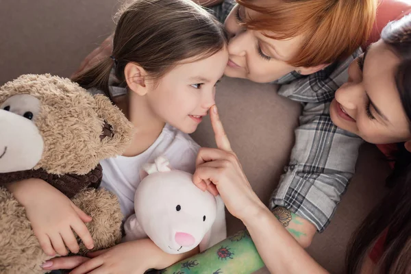 Smiling same sex parents kissing and touching nose of daughter with soft toys on couch — Stock Photo