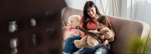 Enfoque selectivo de la madre sonriente mirando a la hija jugando con el oso de peluche en el sofá, plano panorámico - foto de stock