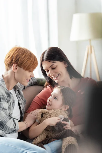 Selective focus of smiling mothers looking at daughter with teddy bear on couch at home — Stock Photo