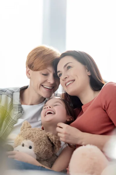Selektiver Fokus des Kindes mit Teddybär, das neben lächelnden gleichgeschlechtlichen Eltern zu Hause lacht — Stockfoto