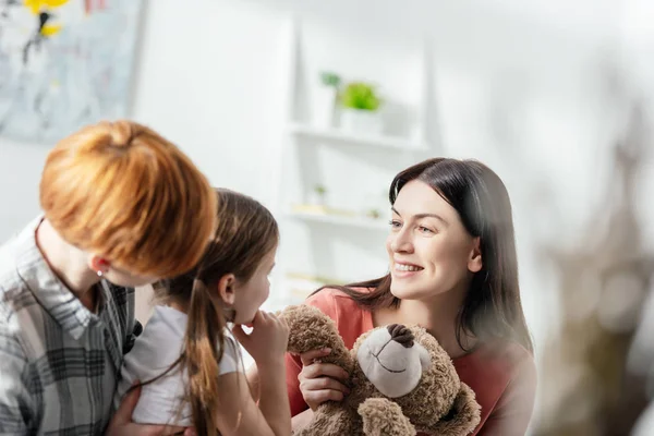 Foco seletivo de mãe sorridente segurando ursinho de pelúcia perto da filha e dos pais na sala de estar — Fotografia de Stock
