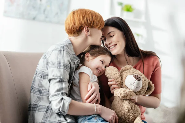 Des mères heureuses embrassant leur fille souriante avec un ours en peluche sur le canapé — Photo de stock