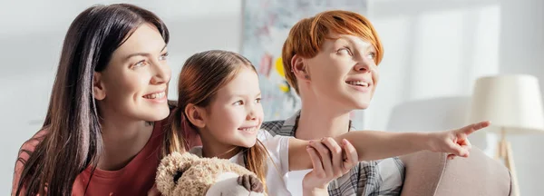 Panoramic shot of smiling kid with teddy bear pointing with finger near mothers on couch — Stock Photo