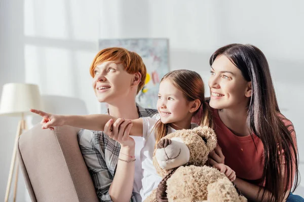 Lächelndes Kind mit Teddybär zeigt mit Finger in die Nähe von Müttern, während es im Wohnzimmer auf der Couch sitzt — Stockfoto