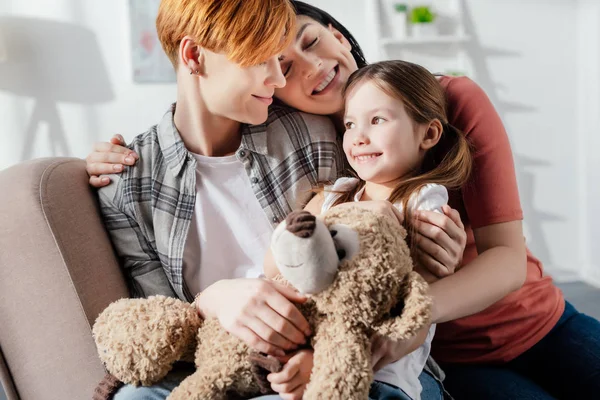 Sonriente familia del mismo sexo abrazando hija con osito de peluche en el sofá - foto de stock