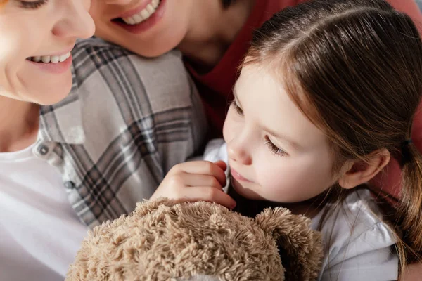 Child with teddy bear looking away near smiling same sex parents — Stock Photo