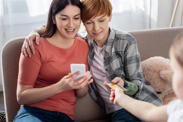 Selective focus of mothers using smartphone and daughter giving credit card in living room — Stock Photo