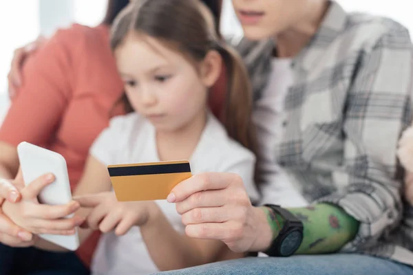 Вибірковий фокус одностатевих батьків, які тримають кредитну картку під час дочки, використовуючи смартфон вдома — стокове фото