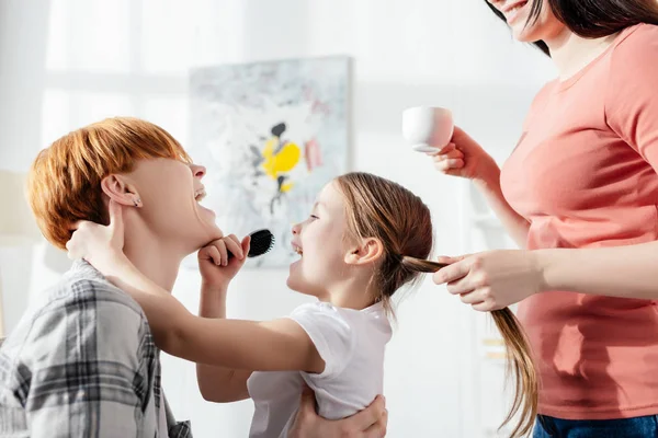 Vue latérale de souriants parents de même sexe riant avec fille tenant peigne dans le salon — Photo de stock