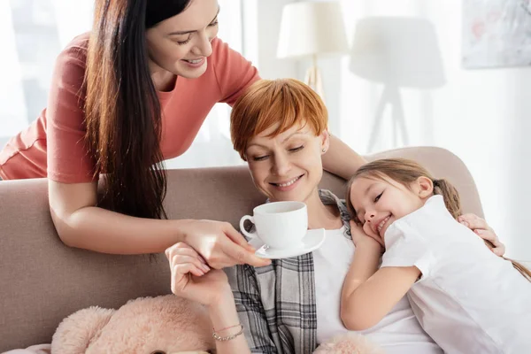 Smiling woman giving coffee to parent hugging happy daughter on sofa — Stock Photo