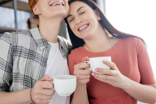 Sonriendo cupé del mismo sexo sosteniendo tazas de café en casa - foto de stock