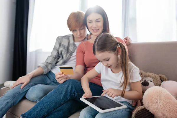 Selektiver Fokus des Kindes mit digitalem Tablet in der Nähe von Müttern mit Kreditkarte auf der Couch im Wohnzimmer — Stockfoto