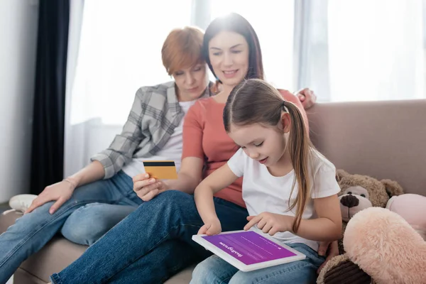 KYIV, UKRAINE - FEBRUARY 10, 2020: Child using digital tablet with instagram app on screen near smiling mothers with credit card on sofa — Stock Photo