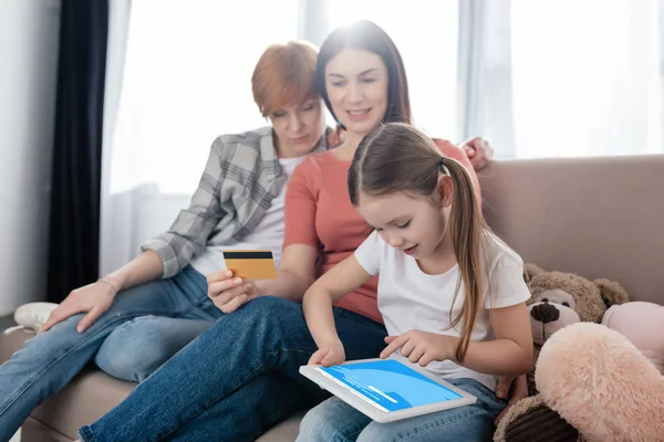 KYIV, UKRAINE - FEBRUARY 10, 2020: Child using digital tablet with twitter app on screen near smiling same sex parents with credit card on sofa — Stock Photo