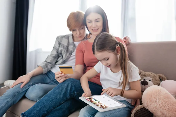 KYIV, UKRAINE - FEBRUARY 10, 2020: Child using digital tablet with foursquare app on screen near mothers with credit card on sofa at home — стокове фото