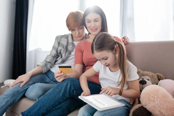 KYIV, UKRAINE - FEBRUARY 10, 2020: Child using digital tablet with vk app on screen near same sex parents with credit card on couch — Stockfoto