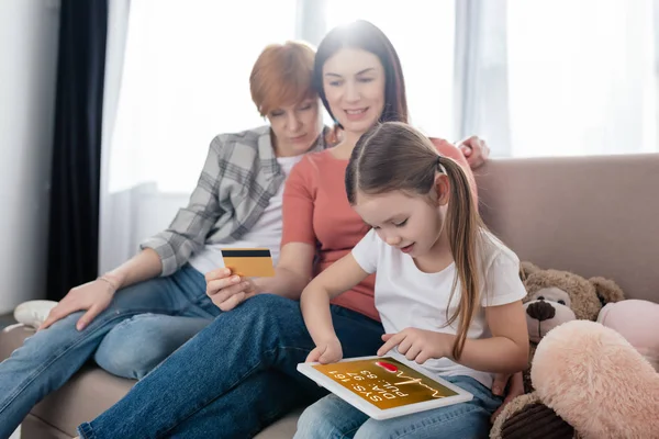 Niño usando tableta digital con aplicación de salud electrónica cerca de sonreír padres del mismo sexo con tarjeta de crédito en el sofá — Stock Photo