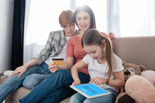 KYIV, UKRAINE - FEBRUARY 10, 2020: Child using digital tablet with skype app on screen near mothers with credit card  on couch — Stockfoto