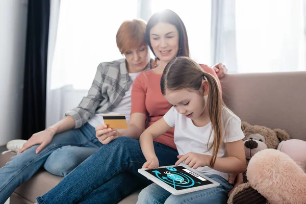 KYIV, UKRAINE - 10 FÉVRIER 2020 : Enfant utilisant une tablette numérique avec application youtube à l'écran près des mères avec carte de crédit sur le canapé — Photo de stock