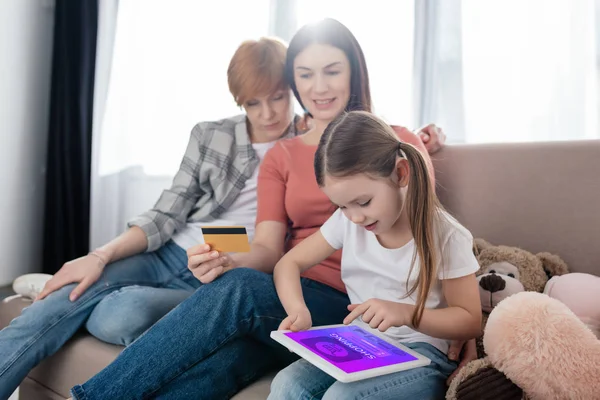 Criança usando tablet digital com aplicativo de compras na tela perto de pais do mesmo sexo com cartão de crédito e brinquedos macios no sofá — Fotografia de Stock