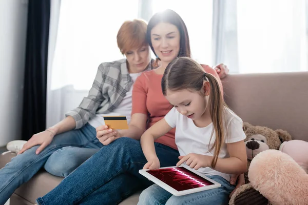 Ребенок, использующий цифровой планшет рядом с матерями с кредитной картой на диване — стоковое фото