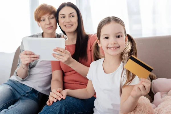 Enfoque selectivo del niño sosteniendo la tarjeta de crédito y sonriendo a la cámara cerca de las madres con tableta digital en el sofá - foto de stock
