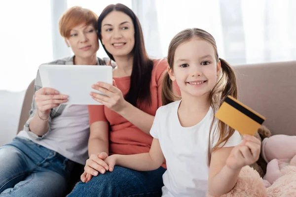Selective focus of kid holding credit card and smiling at camera near mothers with digital tablet on couch — Stock Photo