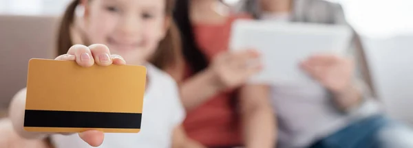 Foco seletivo de criança segurando cartão de crédito e sorrindo para a câmera perto de mães com tablet digital, tiro panorâmico — Fotografia de Stock