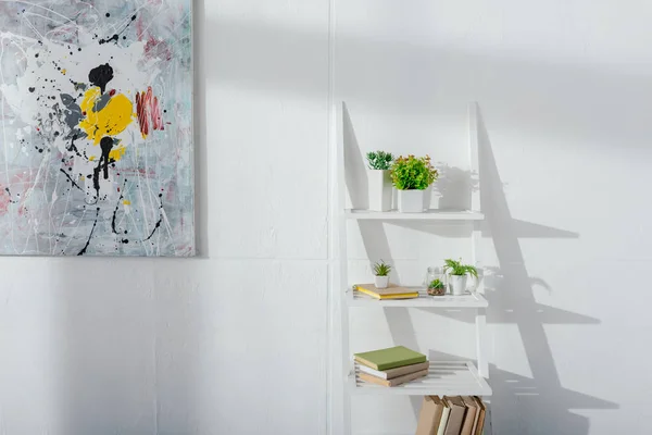 Arte cerca de libros y plantas en la estantería cerca de la pared blanca con luz solar - foto de stock